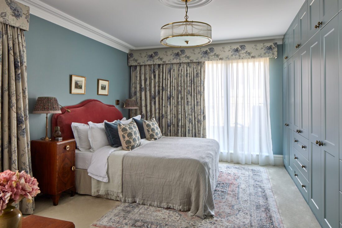 An elegant bedroom designed by Maureen Gomez Interiors, with a large bed featuring an upholstered red headboard, white bedding, and decorative cushions. The room has floral-patterned curtains, Art Deco walnut bedside cabinets, and a rug on the floor. A chandelier hangs from the ceiling.