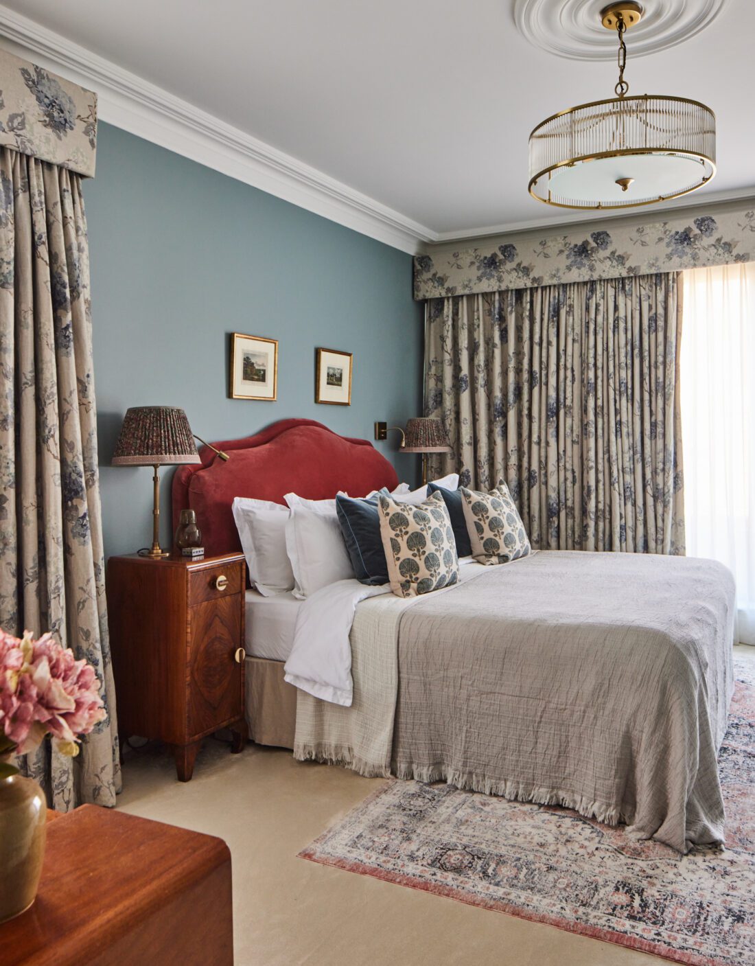 Luxurious bedroom designed by Maureen Gomez Interiors, with a plush bed featuring floral-patterned pillows and a red upholstered headboard. The room has floral curtains, Art Deco nightstands with lamps, and two framed artworks on a teal wall. A patterned rug lies on a cream carpet under a chandelier.