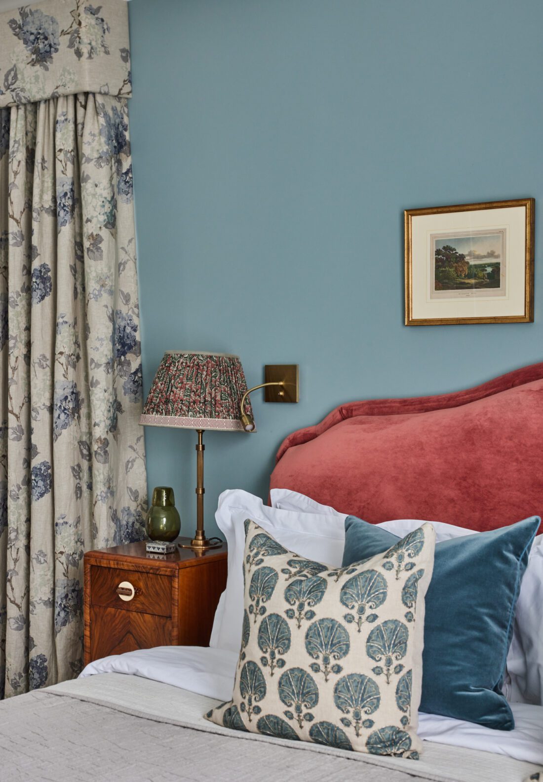 A luxurious bedroom designed by Maureen Gomez Interiors, features a teal wall with a small gold framed picture, a red upholstered headboard, and a walnut Art Deco nightstand. The bed is adorned with patterned and solid coloured pillows. A lamp, books, and a green vase sit on the nightstand beside floral curtains.