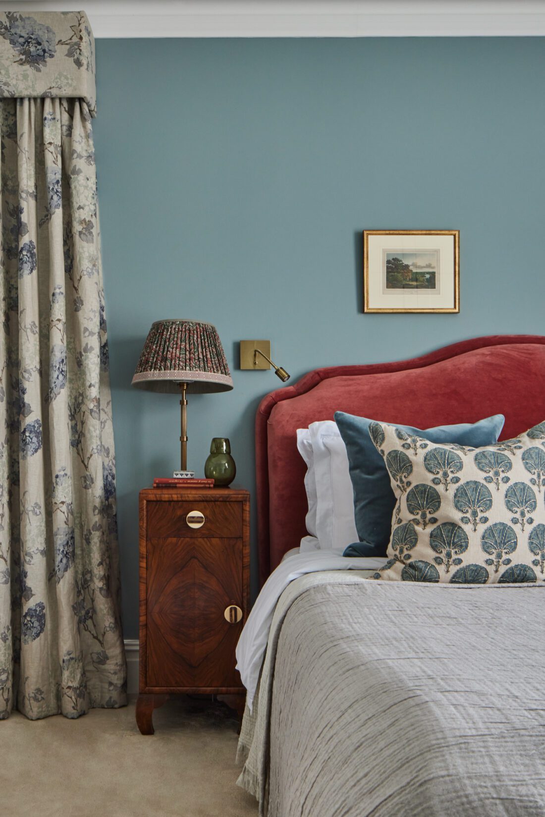 A luxurious bedroom designed by Maureen Gomez Interiors with a red upholstered headboard, teal walls, and floral curtains. An Art-Deco wooden nightstand holds a patterned lamp, books, and a small vase. Decorative pillows with tree prints adorn the bed. A framed picture hangs above.