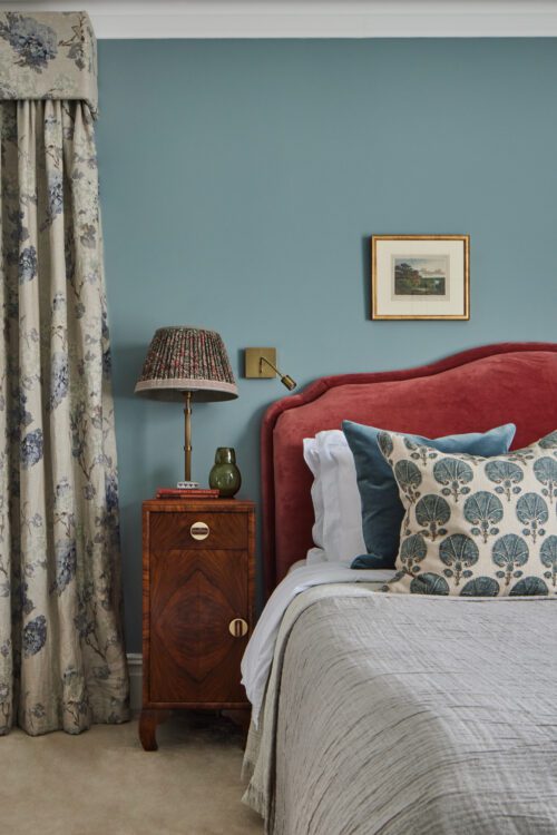 A luxurious bedroom designed by Maureen Gomez Interiors with a red upholstered headboard, teal walls, and floral curtains. An Art-Deco wooden nightstand holds a patterned lamp, books, and a small vase. Decorative pillows with tree prints adorn the bed. A framed picture hangs above.
