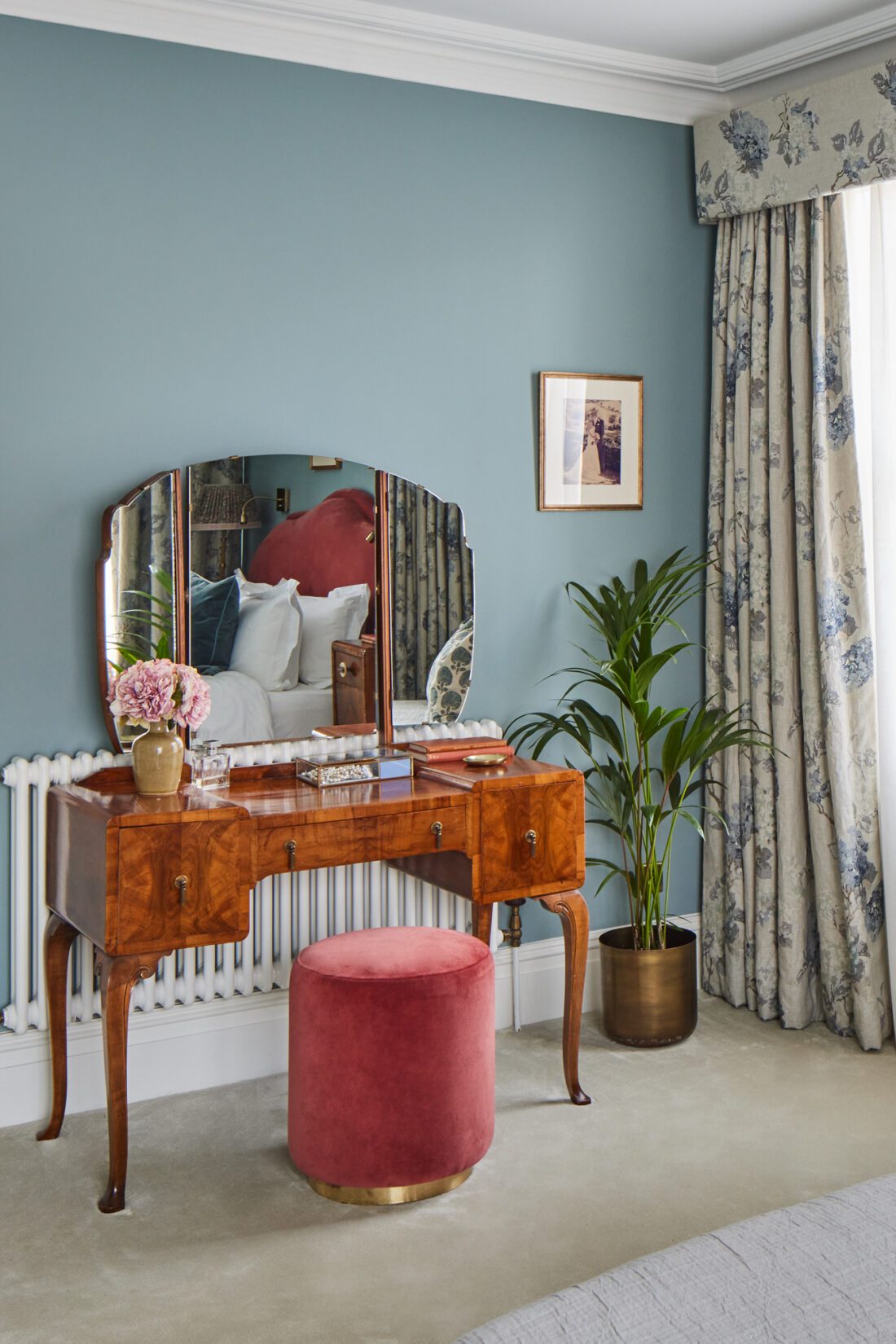 A walnut Art Deco dressing table with a mirror stands against a light blue wall, adorned with flowers and toiletries. A red velvet upholstered stool sits in front. Floral curtains frame a window, and a potted plant adds greenery to this luxurious bedroom scene, designed by Maureen Gomez Interiors.
