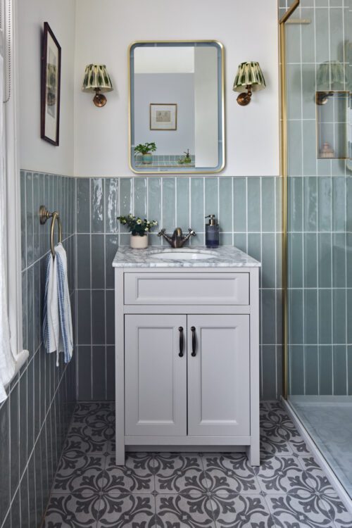 This beautiful bathroom designed by Maureen Gomez Interiors, showcases a white vanity with a marble countertop and chrome tap, adorned with potted plants. A mirror graces the muted green tiled walls, while the patterned grey floor tiles add charm. Above, two wall lights illuminate the serene space.