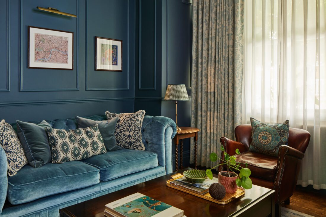An example of interior design in Fulham - An elegant living room with dark blue panelled walls, designed by Maureen Gomez Interiors. It features a brown leather chair, a wooden desk with a computer, an original Victorian fireplace, a turquoise sofa, and a large chandelier. The room is adorned with a potted plant and books on shelves. A window with curtains lets in natural light.