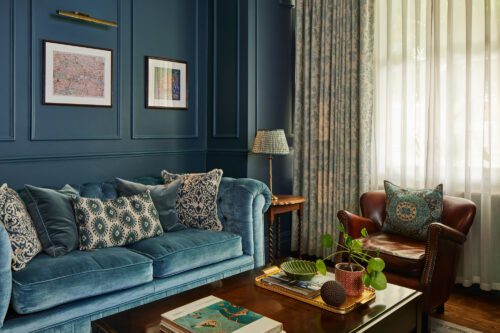 An example of interior design in Fulham - An elegant living room with dark blue panelled walls, designed by Maureen Gomez Interiors. It features a brown leather chair, a wooden desk with a computer, an original Victorian fireplace, a turquoise sofa, and a large chandelier. The room is adorned with a potted plant and books on shelves. A window with curtains lets in natural light.