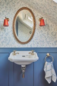 A small bathroom designed by Maureen Gomez Interiors, with delicate blue and white botanical wallpaper and blue panelling. A gold antique oval mirror is centred above a white wall-mounted sink with chrome taps. Orange wall lamps flank the mirror. A blue and white hand towel hangs on a gold towel ring to the right.