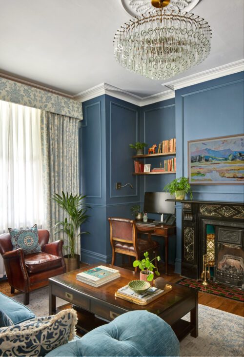 A cosy, elegant living room designed by Maureen Gomez Interiors, with blue walls and a large window with patterned curtains. It features a brown leather chair, a wooden desk with a computer, a fireplace, and a crystal chandelier. The room is decorated with plants and books.