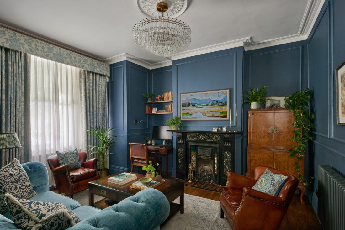 An example of interior design services in Hammersmith - an elegant living room designed by Maureen Gomez Interiors. with dark blue panelled walls, a chandelier, and an original Victorian fireplace. The room features a turquoise sofa, brown leather chairs, a small desk with a computer, bookshelves, and plants. A large painting and patterned curtains add to the decor.