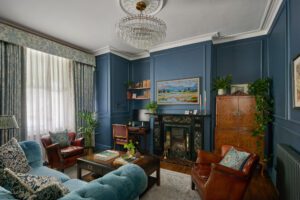 An elegant living room designed by Maureen Gomez Interiors. with dark blue panelled walls, a chandelier, and an original Victorian fireplace. The room features a turquoise sofa, brown leather chairs, a small desk with a computer, bookshelves, and plants. A large painting and patterned curtains add to the decor.