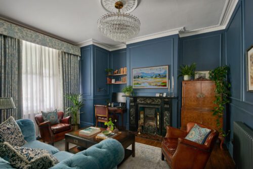 An example of interior design services in Hammersmith - an elegant living room designed by Maureen Gomez Interiors. with dark blue panelled walls, a chandelier, and an original Victorian fireplace. The room features a turquoise sofa, brown leather chairs, a small desk with a computer, bookshelves, and plants. A large painting and patterned curtains add to the decor.