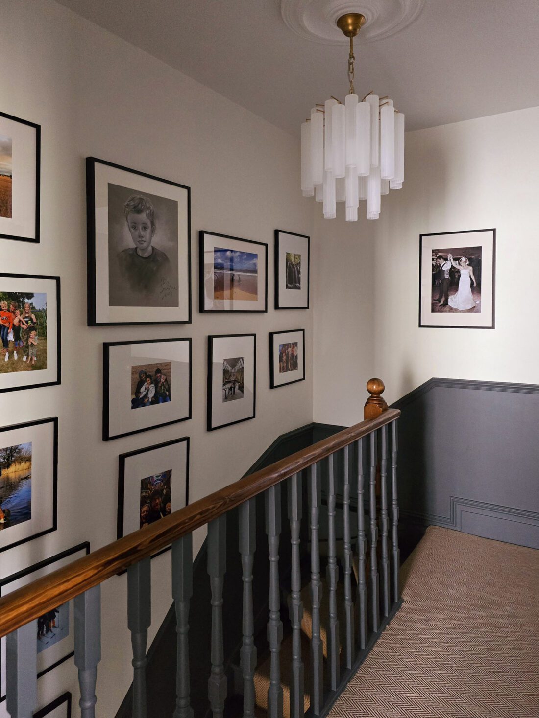 A landing designed by Maureen Gomez Interiors features a staircase with a wooden railing and beige carpet. The walls are adorned with a variety of framed photos. An Art Deco style white, tiered chandelier hangs from the ceiling, and the lower wall panelling is painted grey.