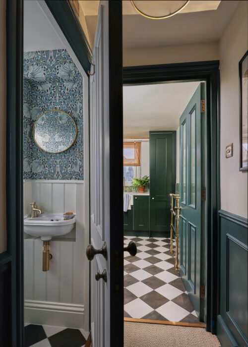 A partially open door reveals two connected rooms: a small bathroom with a floral wallpaper accent and a round mirror, and a larger room with green panelled doors, chequerboard flooring, and a window letting in natural light.