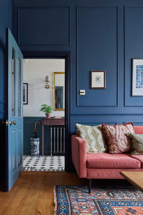 A stylish living room with dark blue panelled walls, a coral sofa with patterned cushions, a colourful rug, and framed artwork. An open door reveals a hallway with black and white tiled flooring and a small table with a plant.
