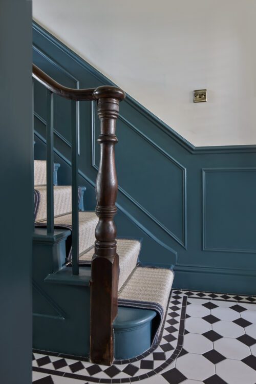 A staircase with dark teal wood panelling, a dark wooden bannister, and beige carpeted steps is shown. The floor has a black and white geometric tile pattern, and a brass light switch is on the wall.