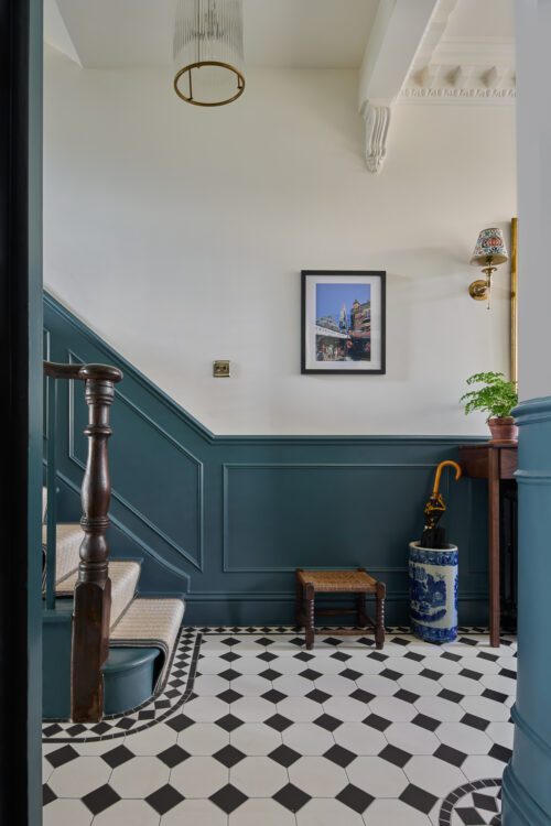 A hallway with black and white chequered floor tiles, teal dado rails, a wooden staircase with beige carpet, a small stool, a framed picture on the wall, and a plant on a console table.