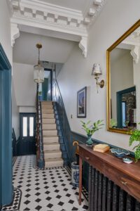 Home 12 A stylish hallway with patterned black-and-white tiled flooring, a carpeted staircase with teal trim, ornate ceiling mouldings, a console table with books and plants, and a large mirror on the wall.