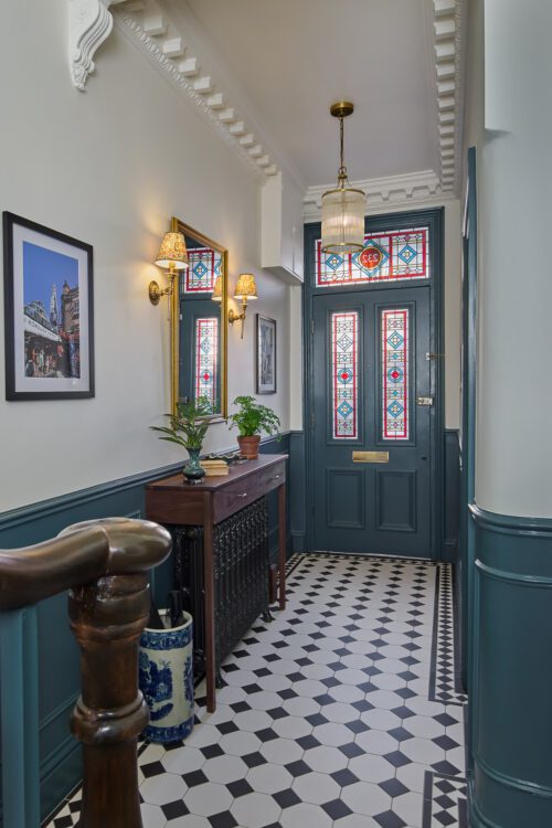 A hallway with a black and white tiled floor, blue dado rail, and a dark blue front door featuring colourful stained glass panels. A wooden table with a lamp, mirror, and potted plants sits against the wall.
