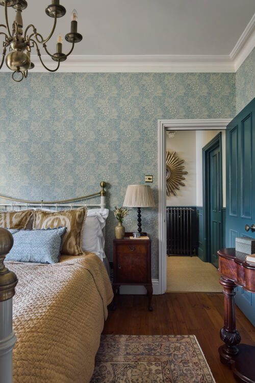 A cosy bedroom with blue patterned wallpaper, a brass bed with gold and blue pillows, a wooden bedside table with a lamp, and a doorway showing a hallway with a decorative sunburst mirror on the wall.