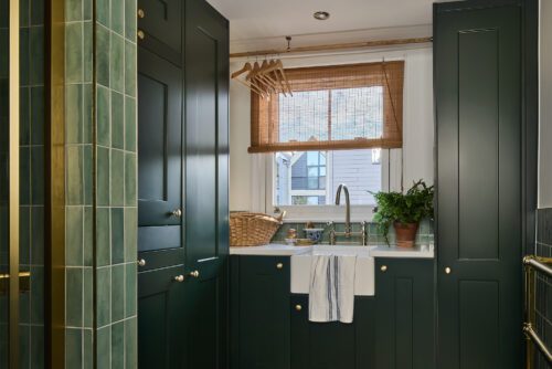 A laundry room with dark green cupboards, a farmhouse sink, a striped towel, a wicker basket, wooden hangers, a potted plant, and a window with a bamboo blind letting in natural light.